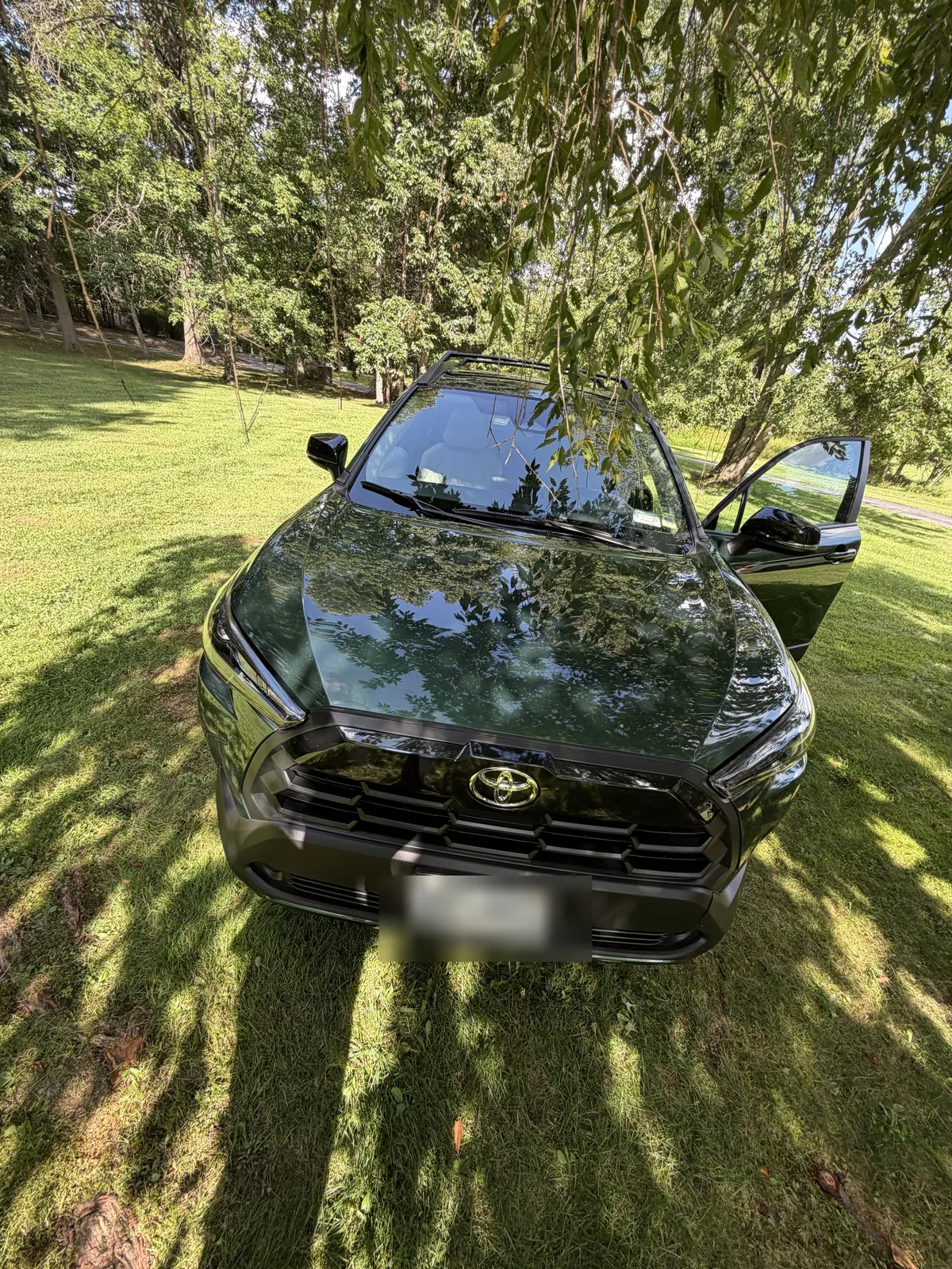 Dark green Toyota Corolla Cross front view on a lawn. The license plate is covered.